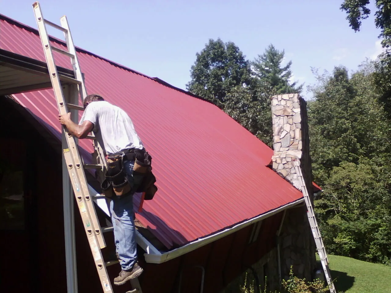 Roofer on a ladder beside a red metal roof and stone chimney — Soffit & Fascia Repair by Roofing Brothers in Taylors Falls